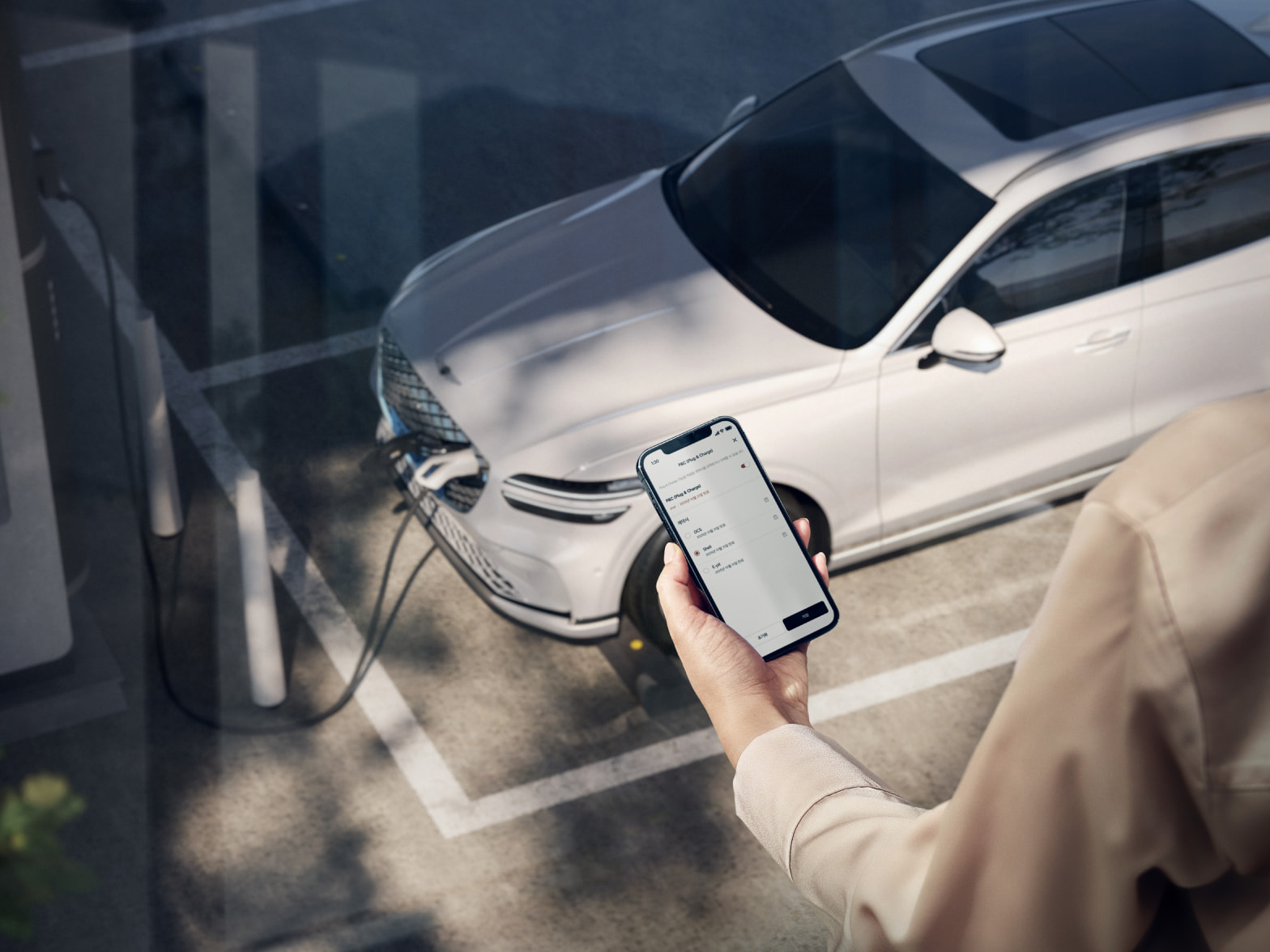 A person is holding a phone while looking down at their vehicle being charged. The vehicle and a arm of person holing a phone are visible together.
