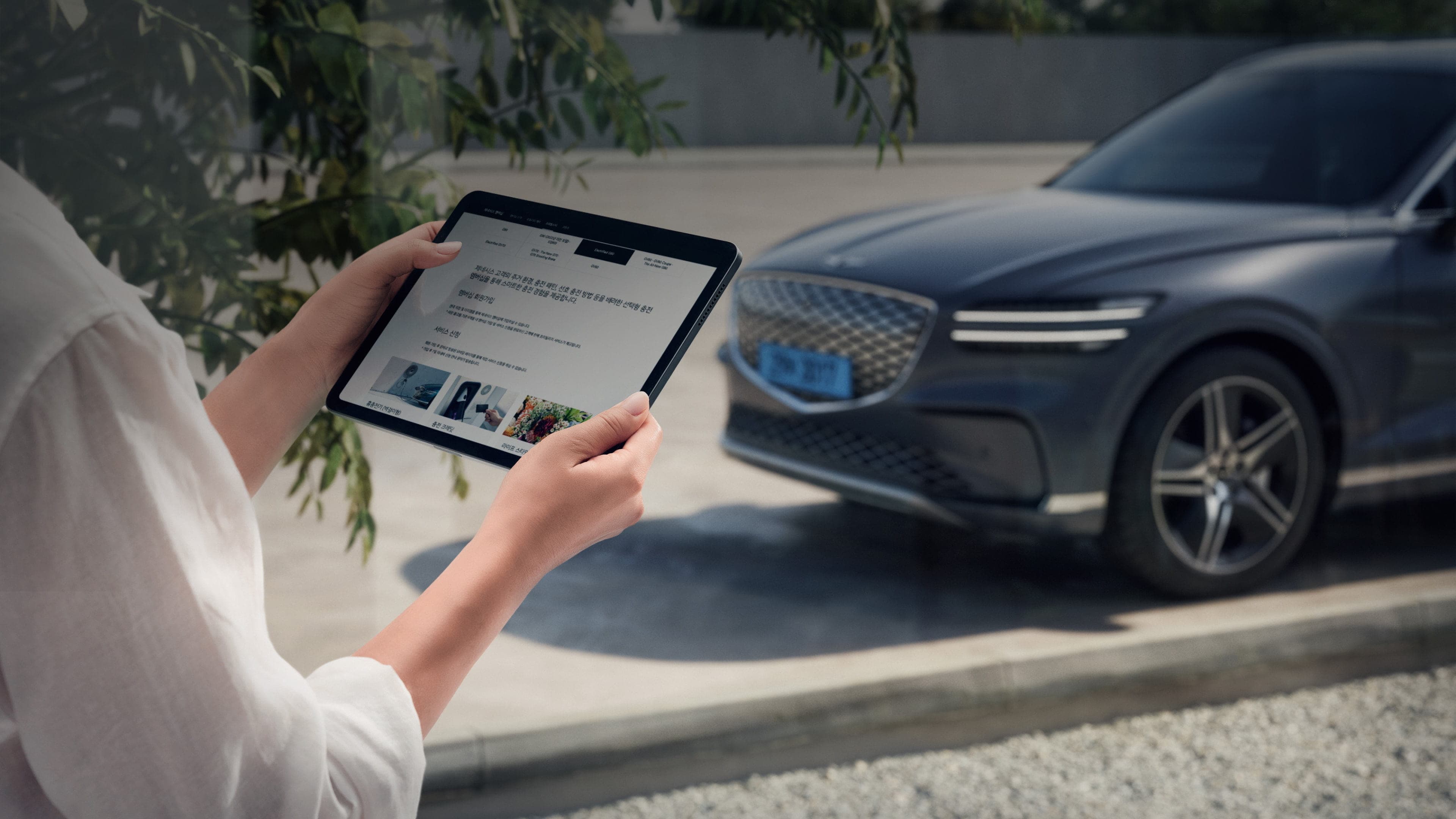 A person holding a tablet PC stands near a parked Genesis vehicle. Parts of the vehicle and the upper body of the person are visible together.