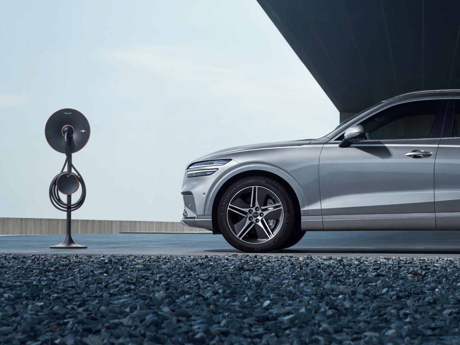 The Electrified GV70 is parked in front of a modern building, with a charger in front of the vehicle.