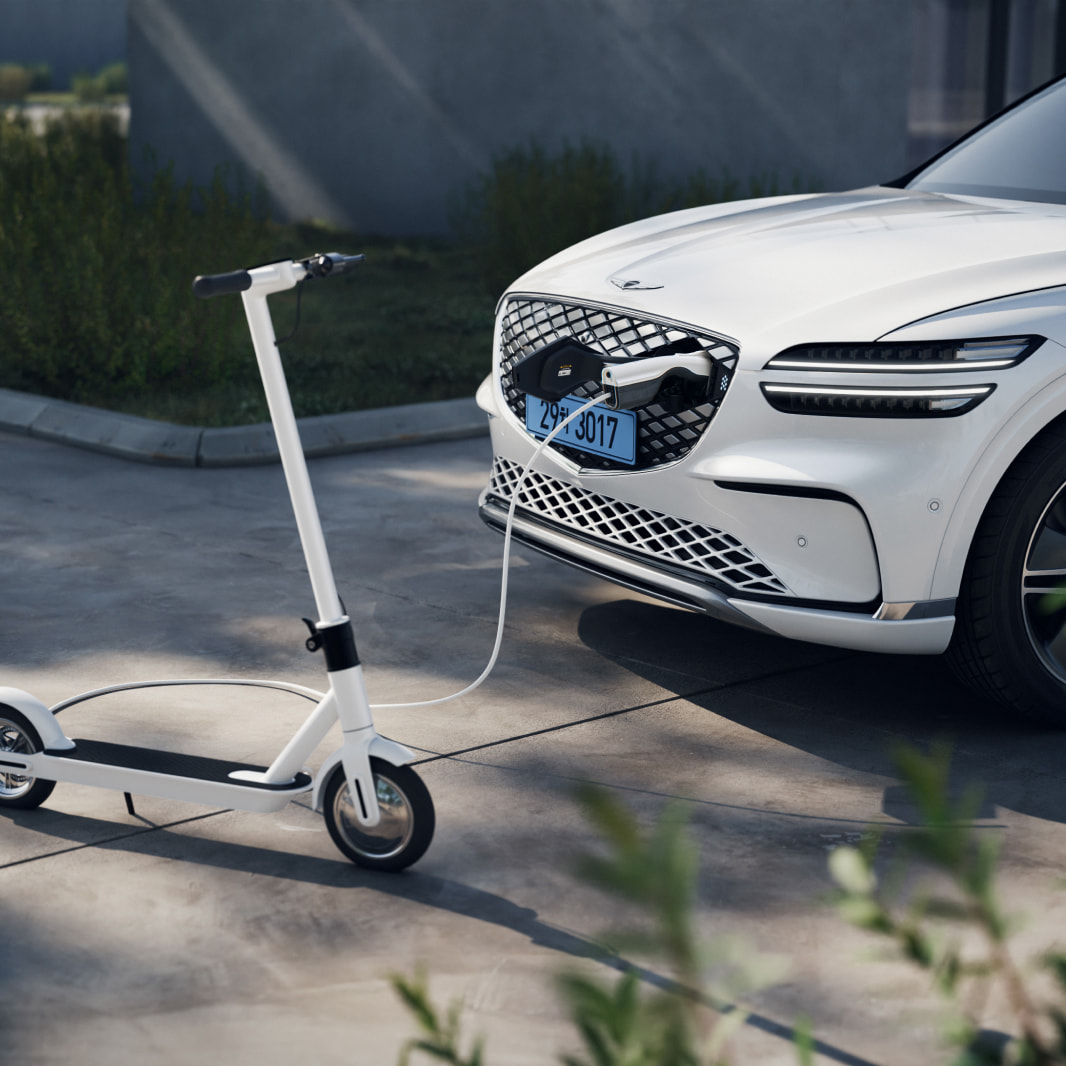 The gender connector plugged into the front charging port of the Genesis electric vehicle is connected to an electric scooter in front of the vehicle.