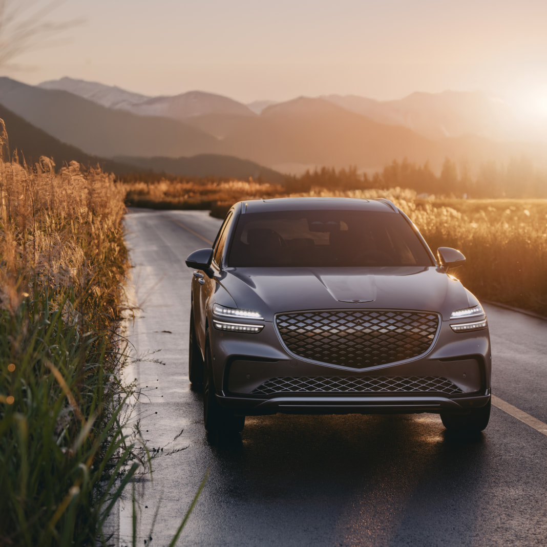 The Electrified GV70 drives along a road surrounded by fields of reeds with the sunset in the background.