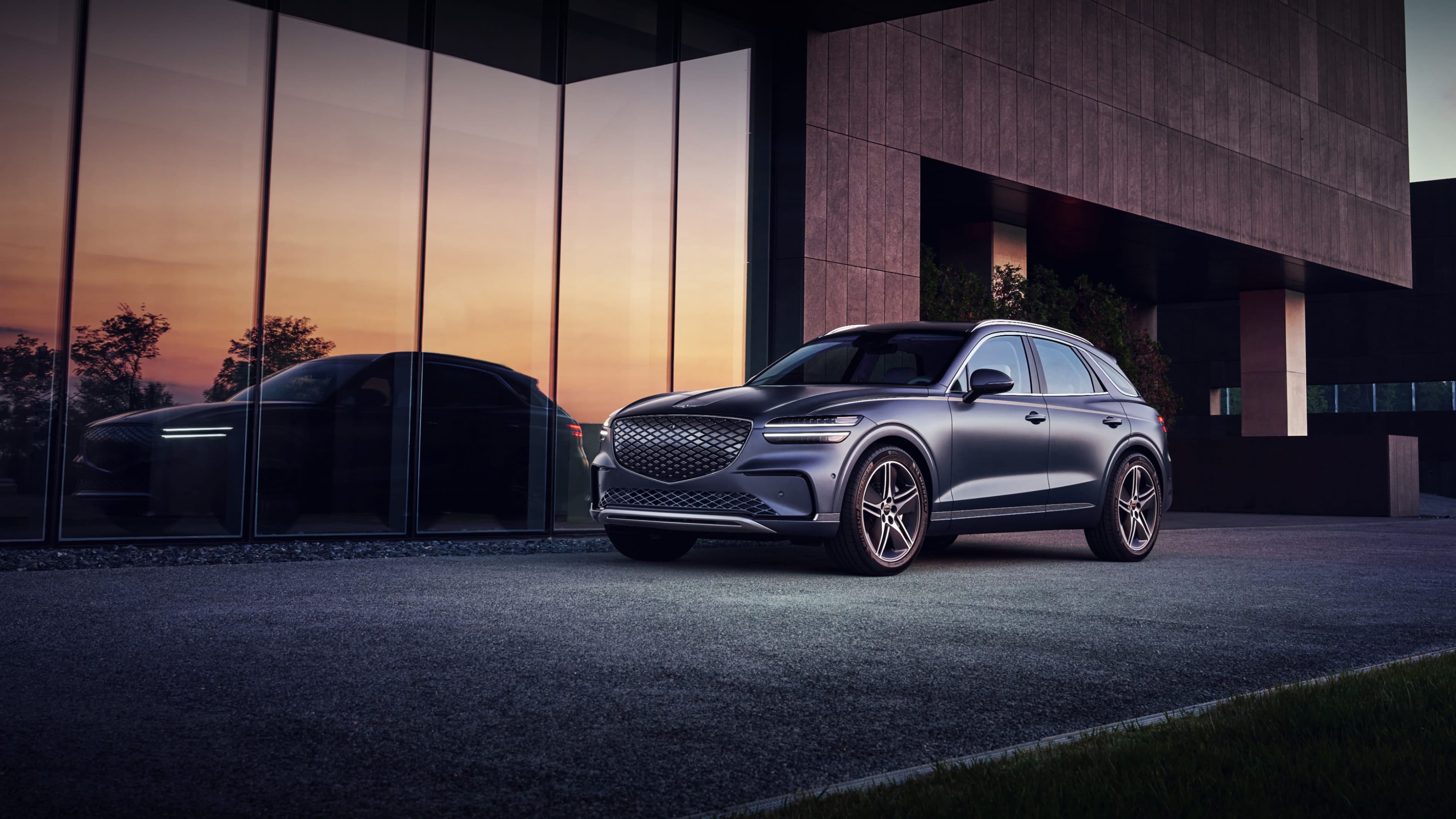 The Ceres Blue Electrified GV70 appears stationary in front of a modern-looking building. The angle provides a view of the vehicle's front and side. The silhouette of the vehicle is the reflected in the windows of the building's first floor.