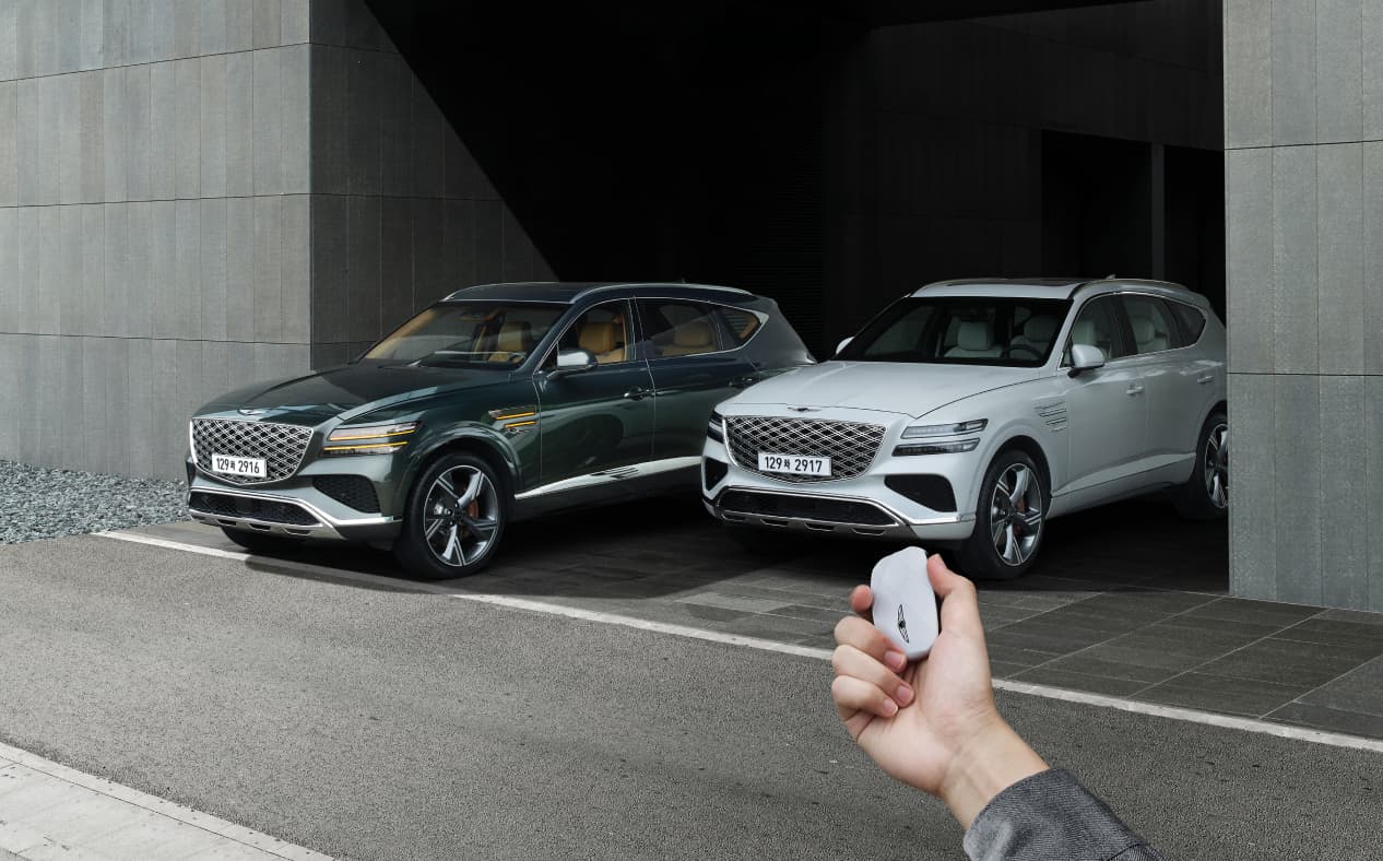 A Vik Black GV80 Coupe and a Uyuni White GV80 Coupe are parked, and the driver is operating the smart key outside the vehicle.
