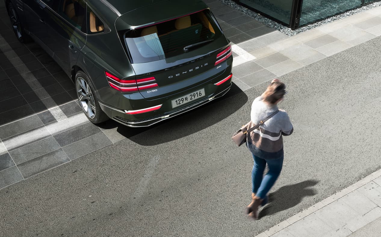 Storr Green GV80 Coupe backing out of a parking lot, and a woman is passing behind the vehicle.