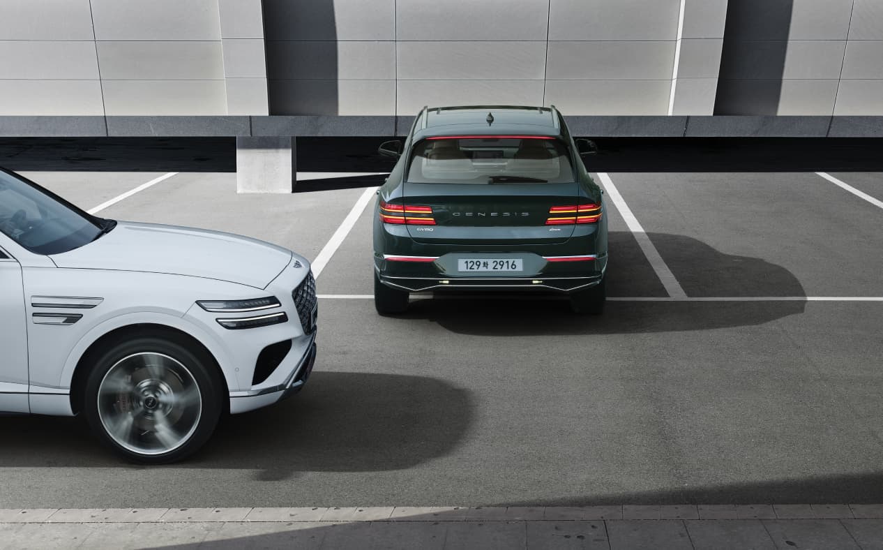 Storr Green GV80 Coupe is backing out of a parking spot, and a Uyuni White GV80 Coupe passes behind the vehicle on the left.
