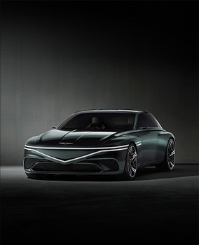 The Genesis X Speedium Coupe Concept vehicle, featuring a dark greenish tint, is parked in a darkened interior. It is bathed in white light, with the front lights turned on. The top row of two front lights extends horizontally, while the bottom row forms a gentle V-shape at the center of the car’s front.