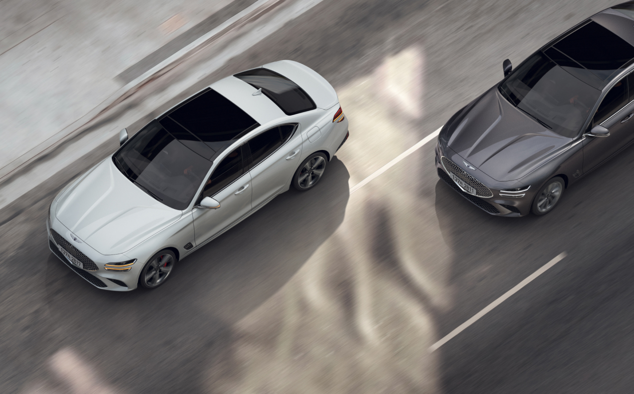 A Makalu Gray G70 is driving toward the rear of a Uyuni White G70 that has turned on its turn signal to change lanes.