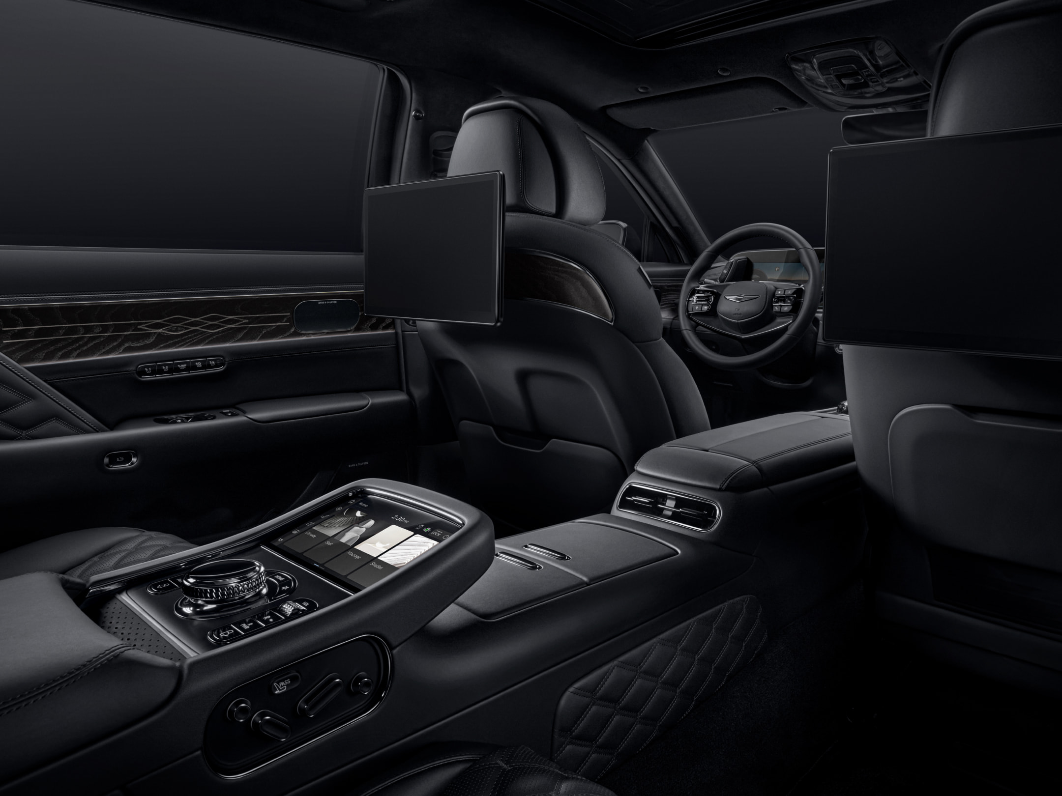 The rear seat interior of the G90 Long Wheel Base Black unfolds from the perspective of the rear seat, looking toward the driver’s seat. The power-operated rear smart entertainment system and center console are visible, with all elements finished in black.