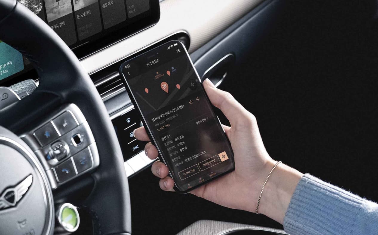 Driver sits in the driver's seat, checking electric charging station information using the My Genesis app.