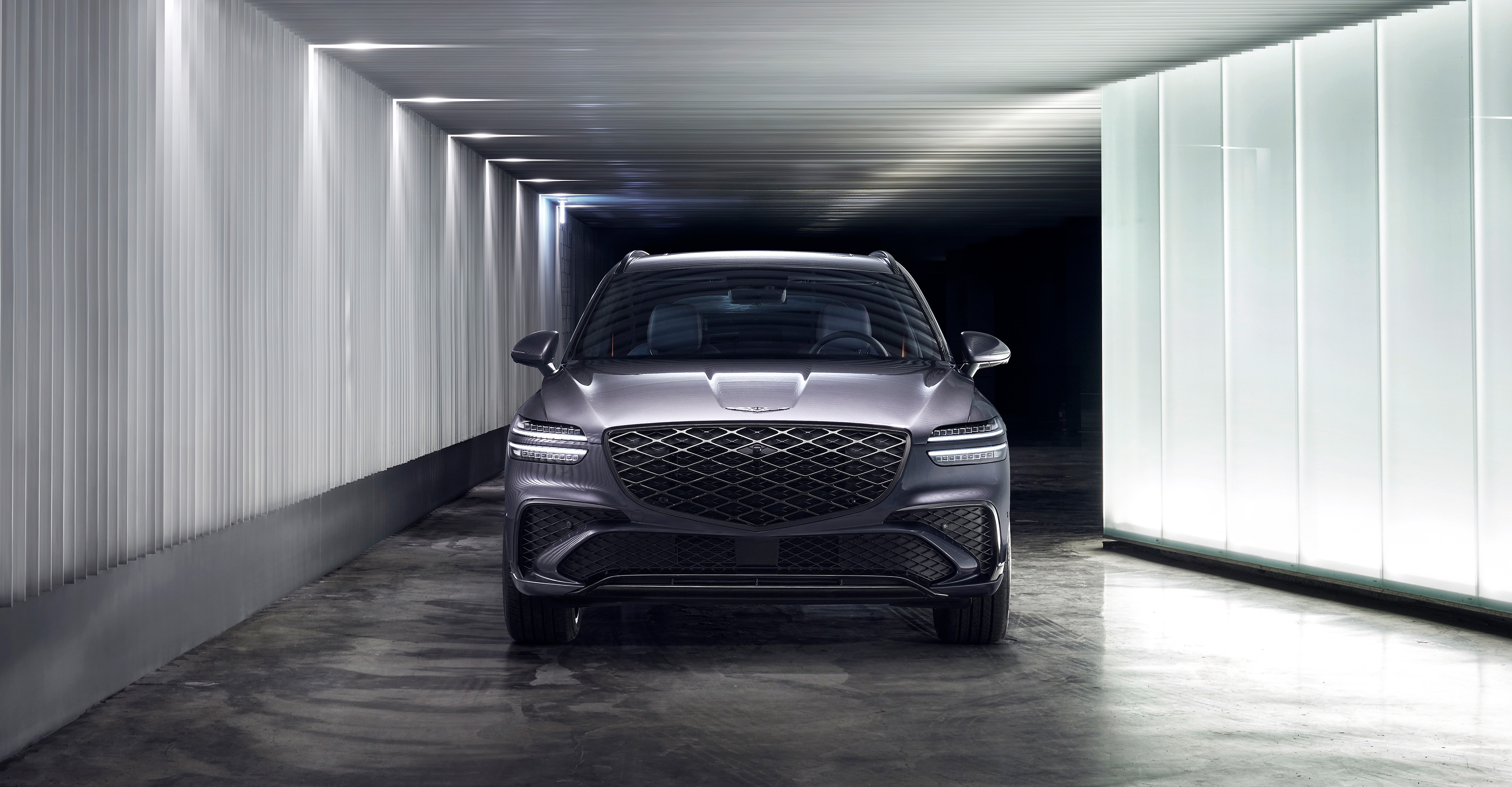 In a dimly lit but brightly illuminated indoor space, a Ceres Blue GV70 sports model is parked, facing forward.