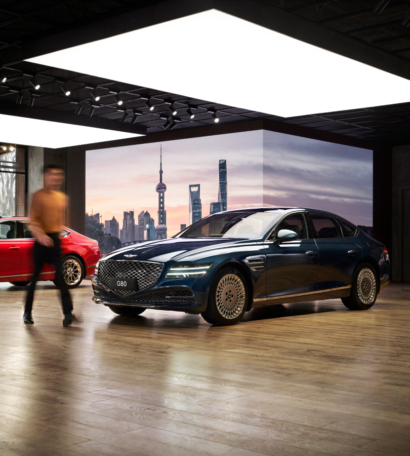 A white Genesis vehicle is on display under wide lighting. Sunlight comes through the window next to it.