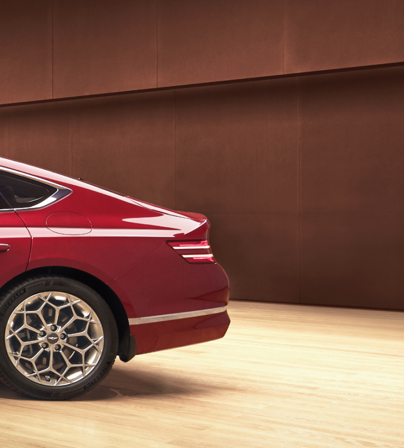 A partial view of the rear end of a red Genesis vehicle. The wall behind it is made with weathered steel.