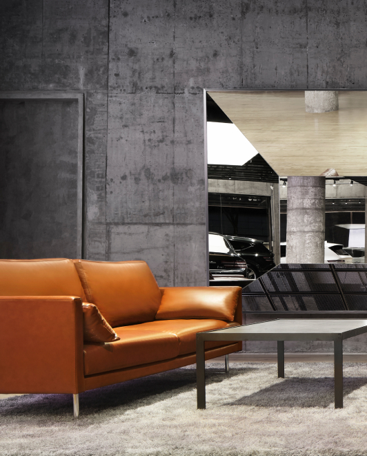 A polyhedral mirror hangs on the wall, with a Genesis vehicle in its reflection. In front of the mirror is a sofa and table.