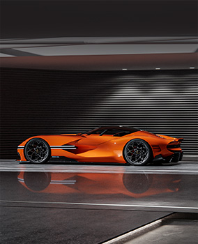 An orange-hued Genesis X Gran Berlineta Concept vehicle is displayed indoors, illuminated by white lights that highlight its low and sleek sports car body. The metallic interior flooring distinctly reflects the vehicle's body.